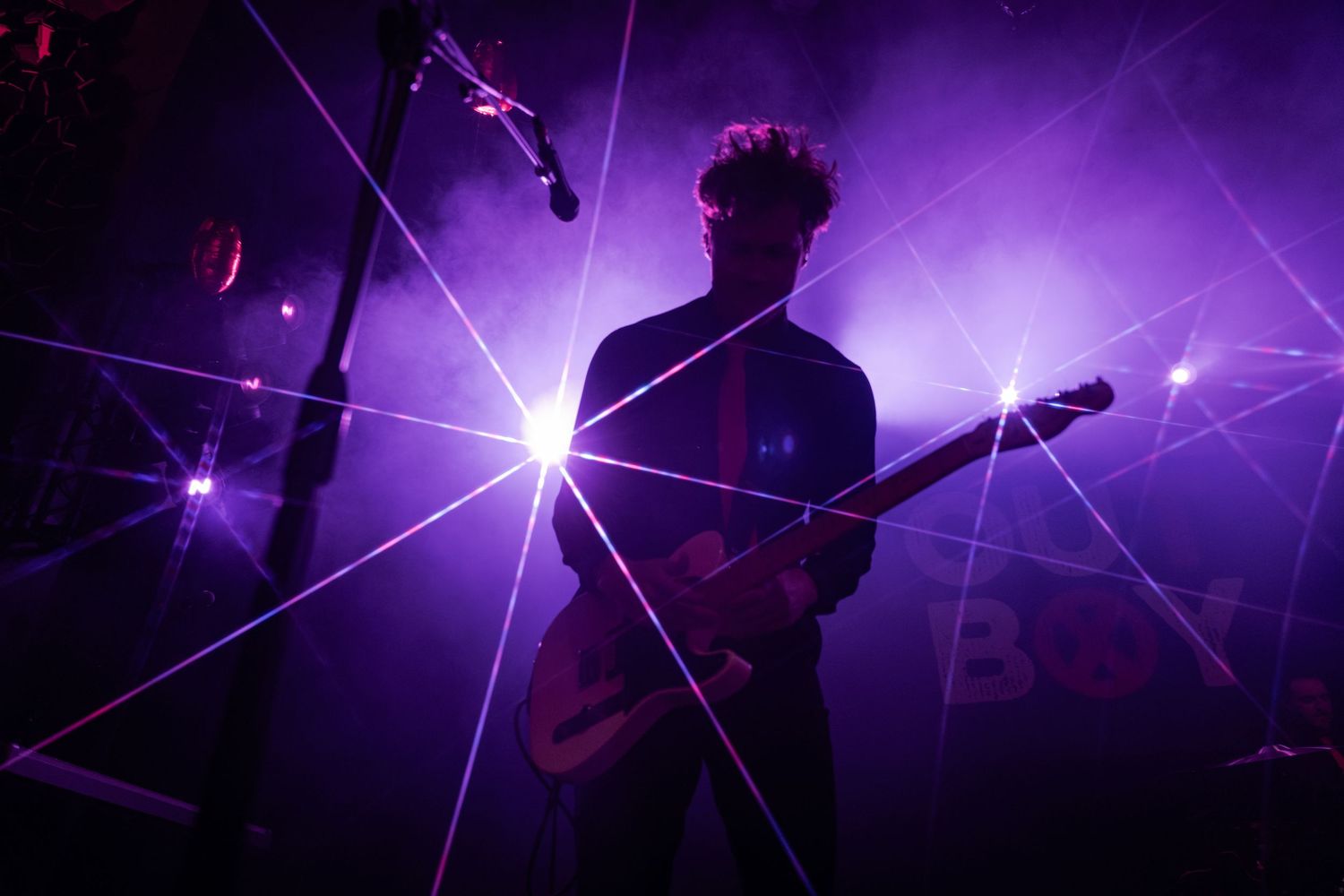 Silhouetted guitarist performing on stage surrounded by vibrant purple laser lights and smoke at a live concert.
