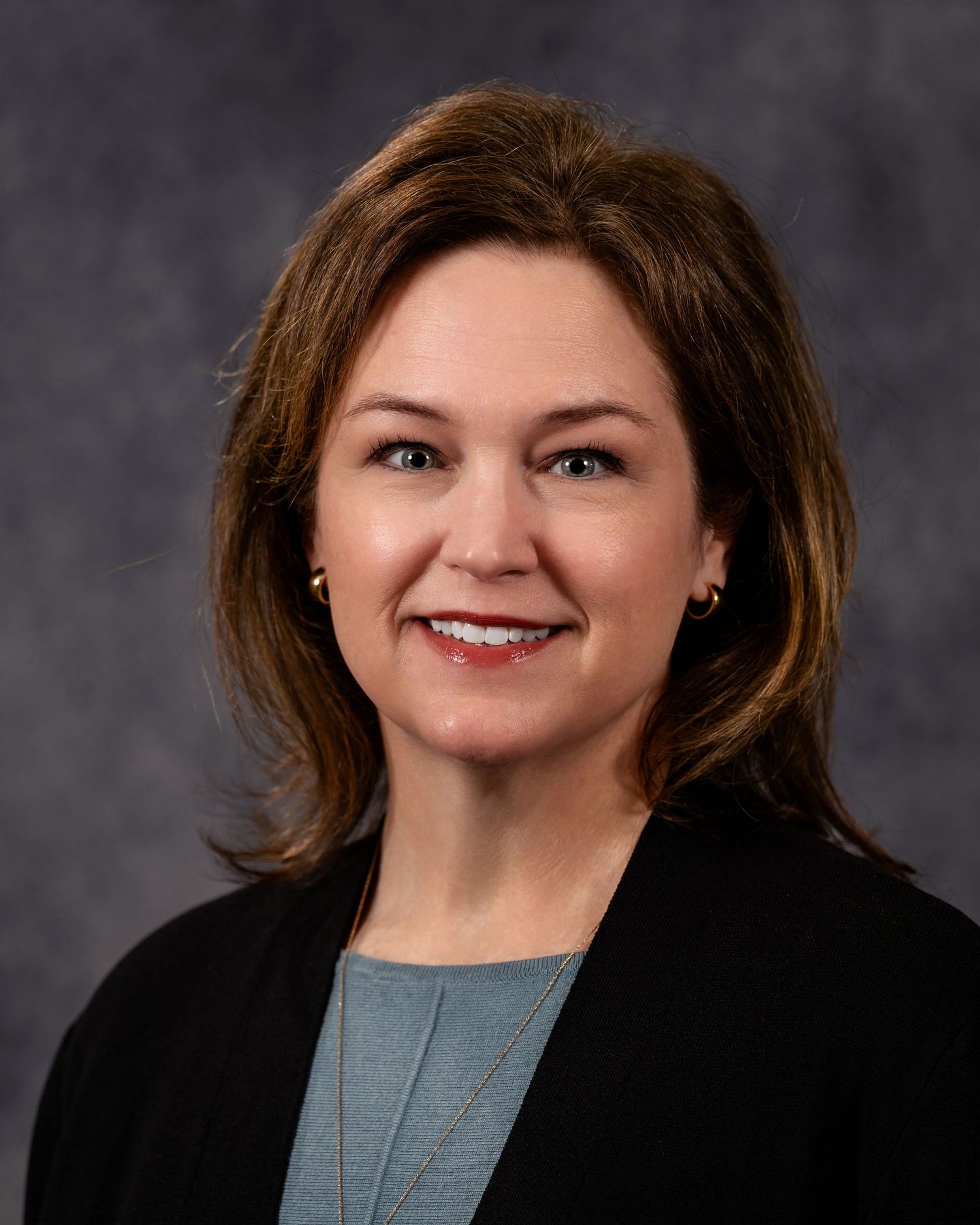 Professional headshot of a smiling woman with brown hair wearing a black blazer and teal top against a gray background.
