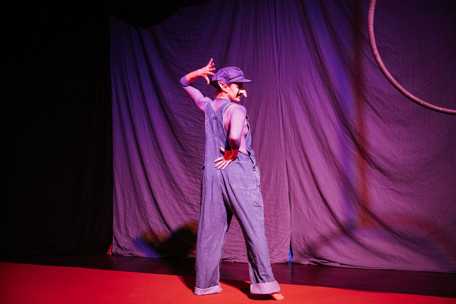 A performer in purple overalls and cap poses dramatically on a red stage floor before a purple draped backdrop.