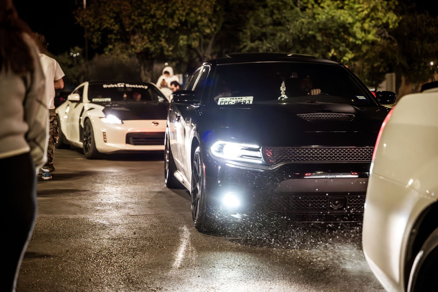 Modified performance cars with LED lighting cruising through a nighttime car meet.