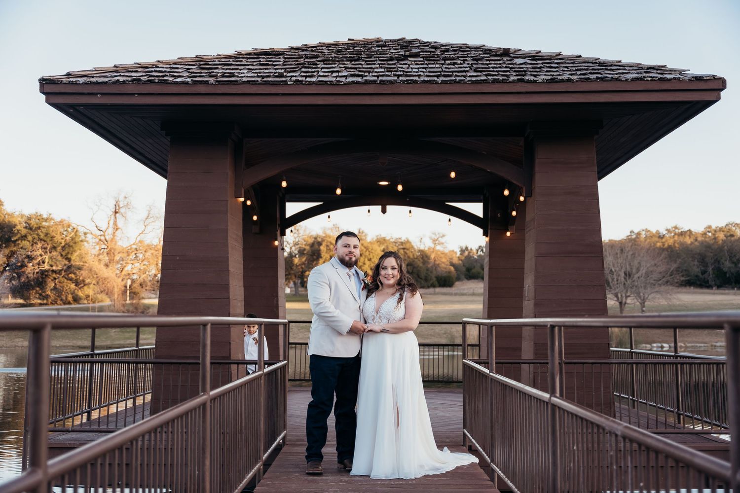 A couple stands on a wooden bridge under a covered pavilion at sunset.