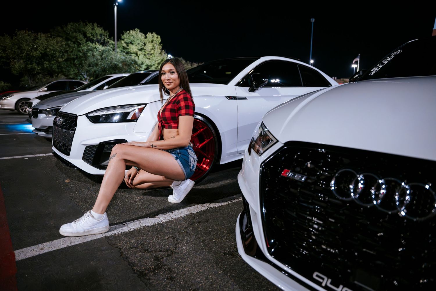 Model in red plaid top poses with white performance cars at night car meet.