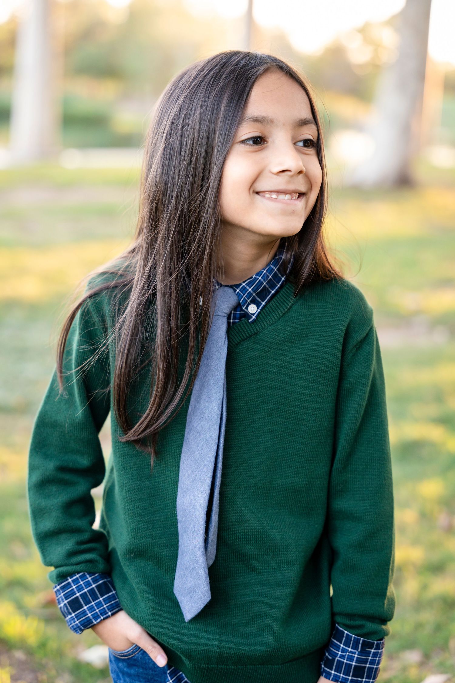Person in green sweater and plaid shirt with gray tie smiling outdoors.