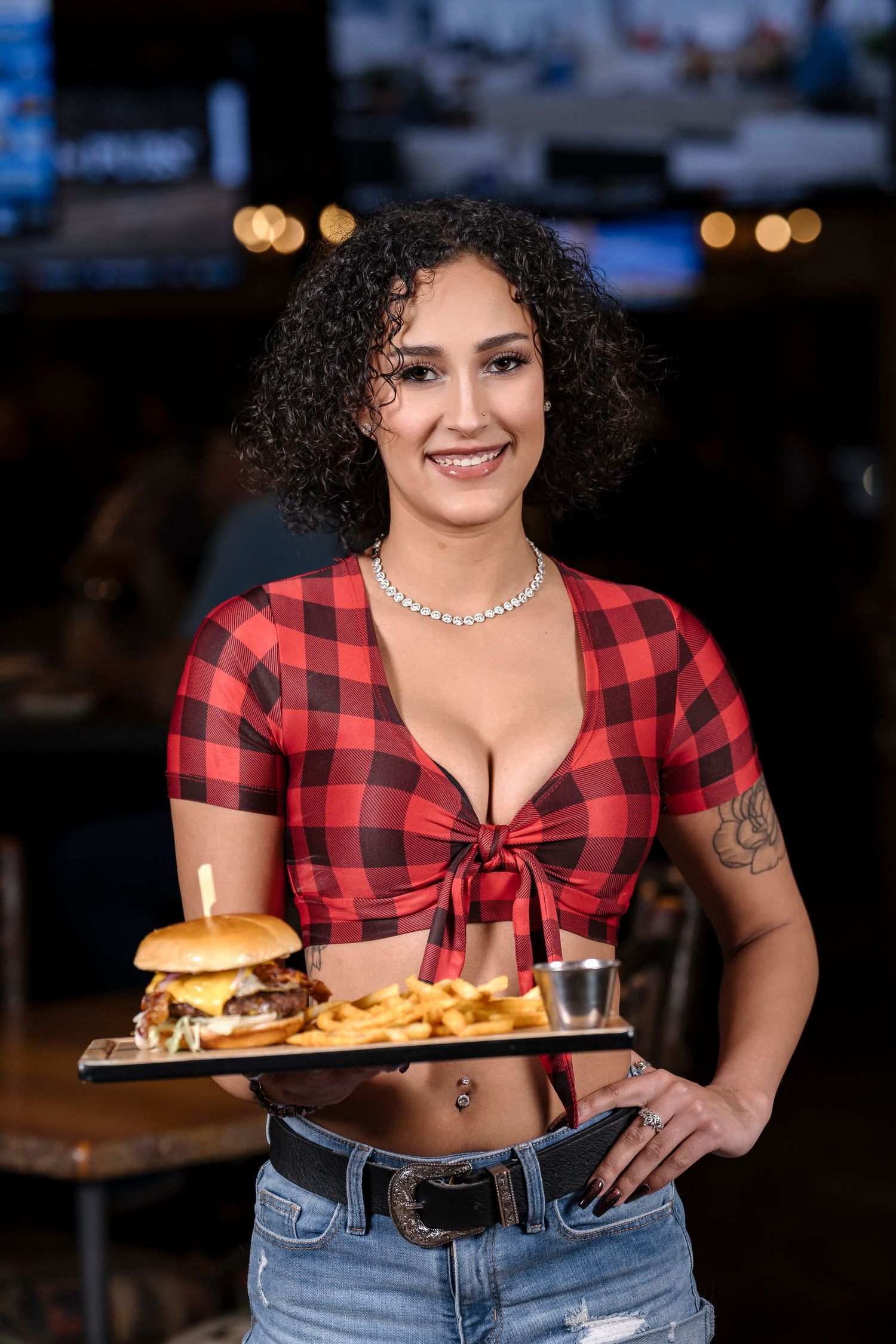 Server in buffalo plaid crop top presents hamburger and fries on wooden serving board.