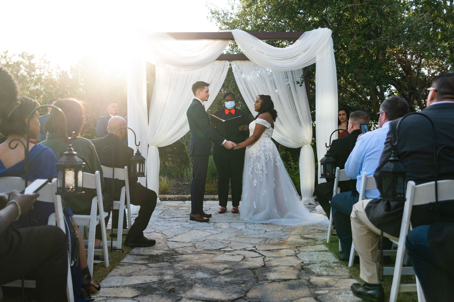 Outdoor wedding ceremony under white draped arch during sunset.