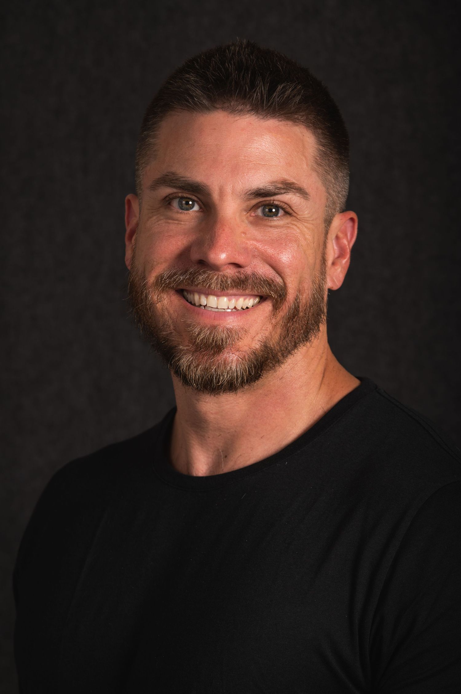 Professional headshot of a person in black t-shirt with neat beard against dark background.