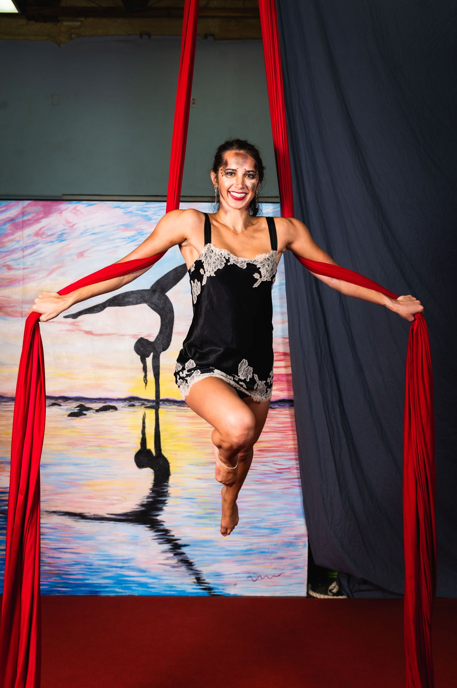 Performer gracefully poses in red aerial silks against painted sunset backdrop.