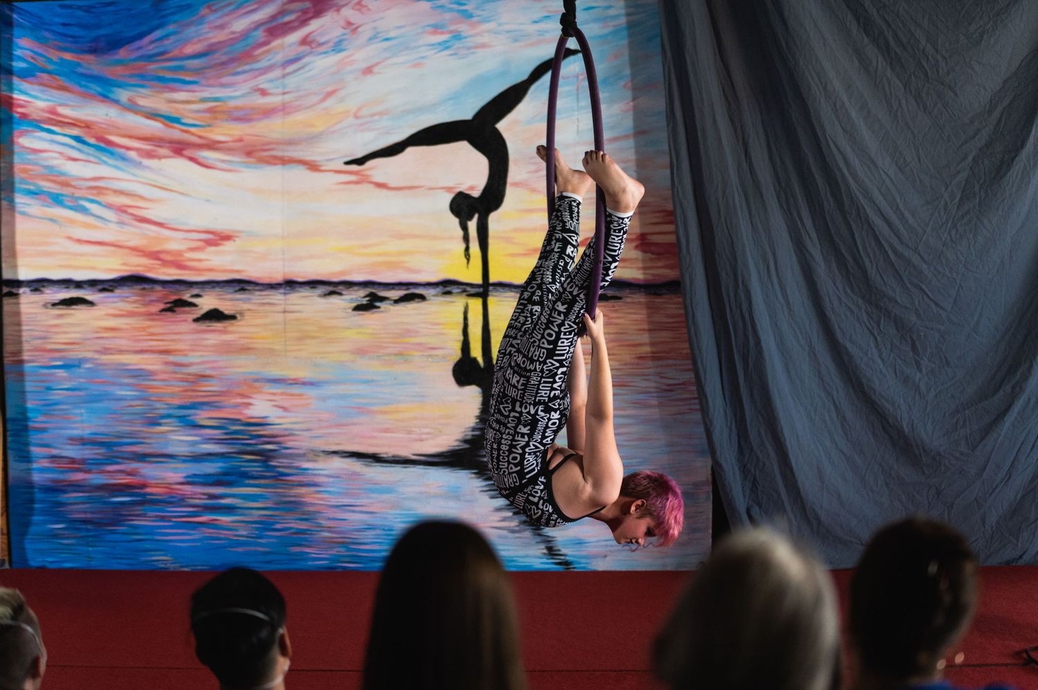 Aerial artist performs graceful pose against vibrant painted sunset backdrop with audience silhouettes.