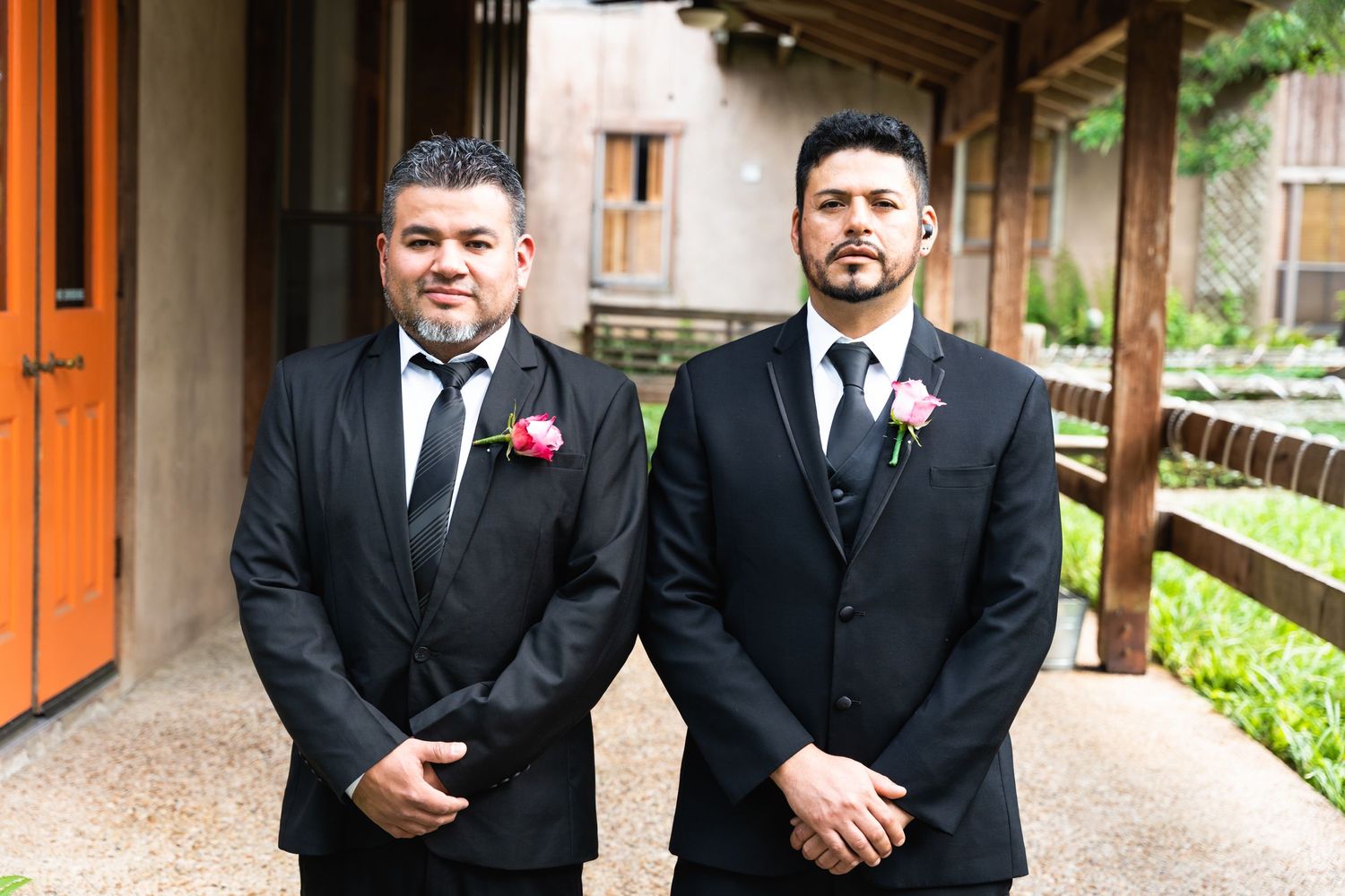 Two groomsmen in black suits with pink boutonnieres stand on a covered porch.