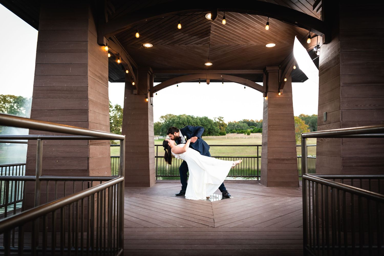 A romantic dip pose captured under a wooden gazebo overlooking a scenic golf course at sunset.