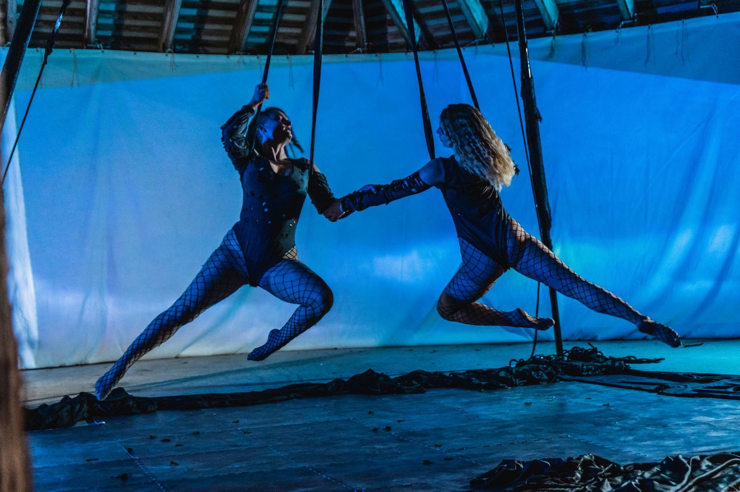 Two performers execute synchronized aerial dance moves against bright blue background.