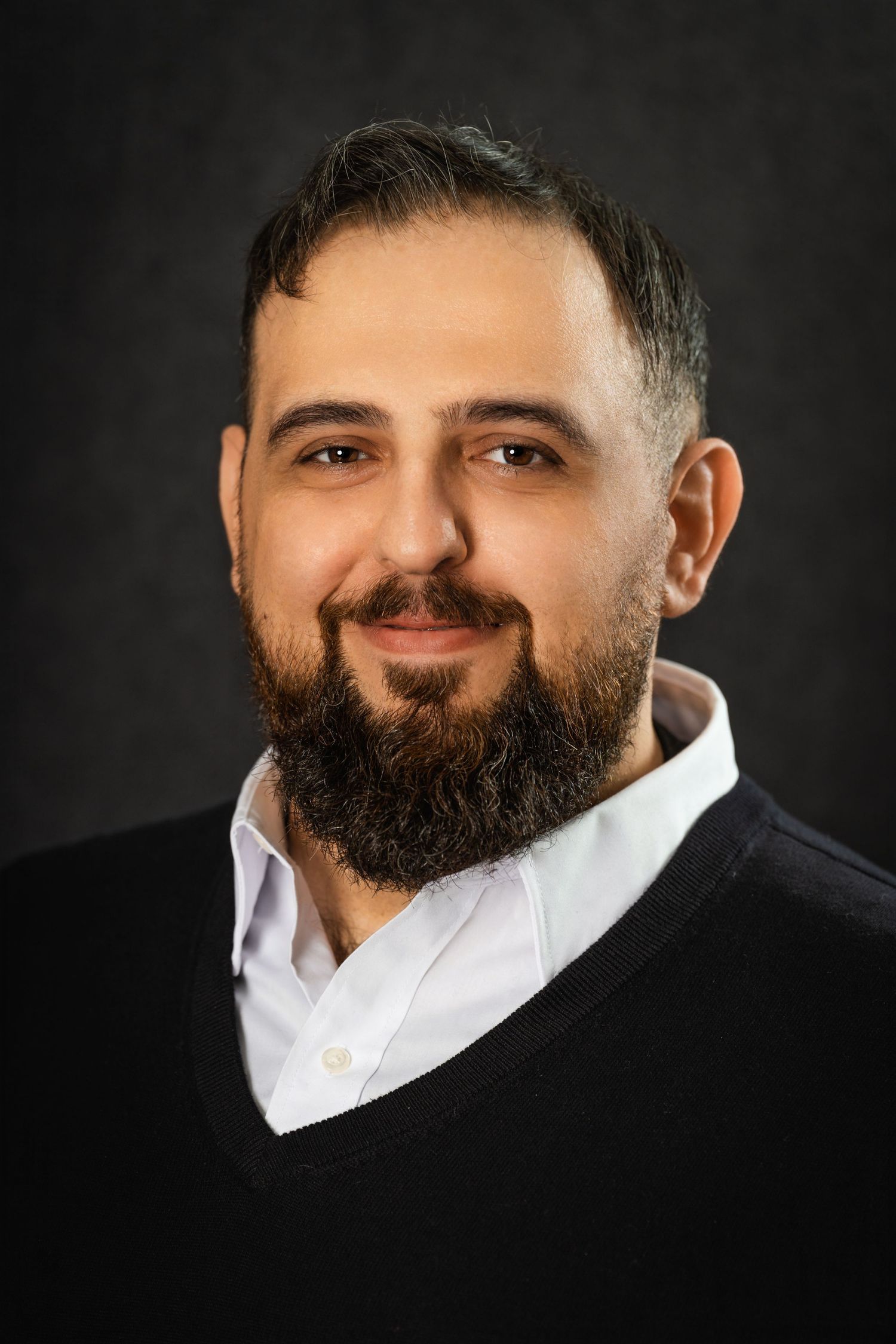 Professional headshot of person in black sweater and white shirt against dark background.