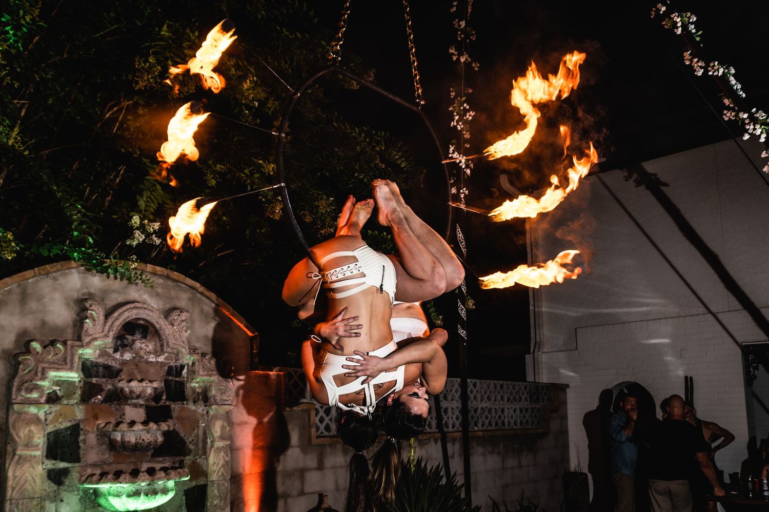 Aerial fire performer suspended in dramatic pose surrounded by flames at night.