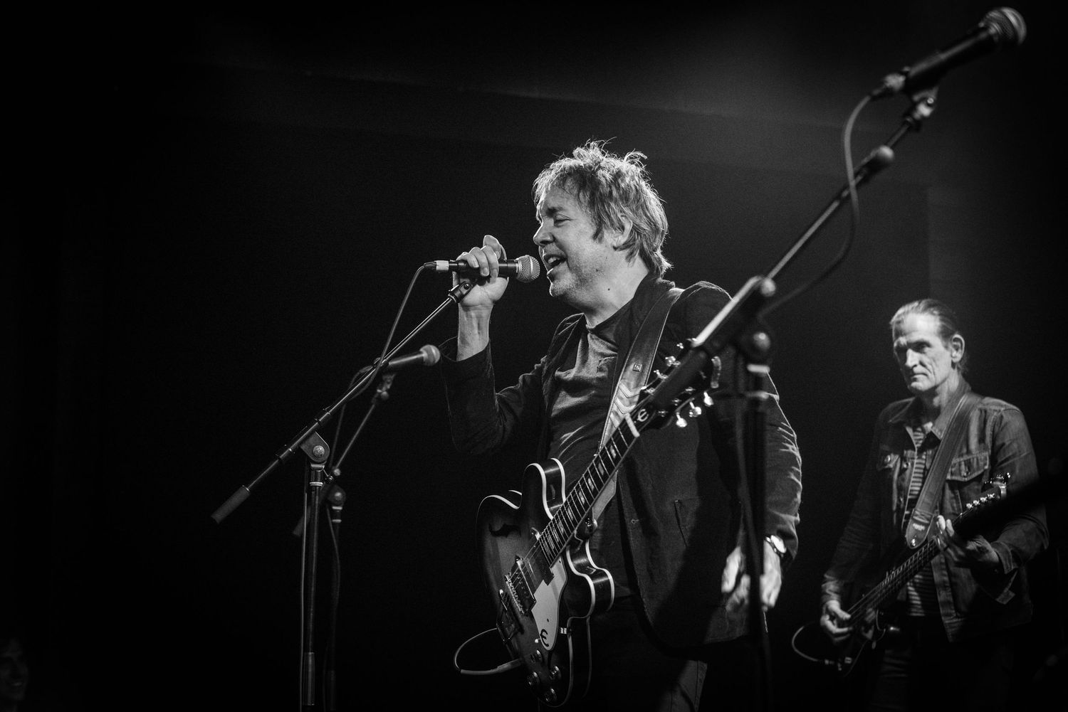 Black and white concert photo of musicians performing with guitars and microphone setup.