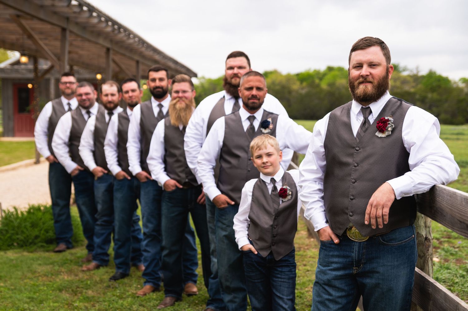 A wedding party in matching grey vests and jeans pose in a line outdoors.
