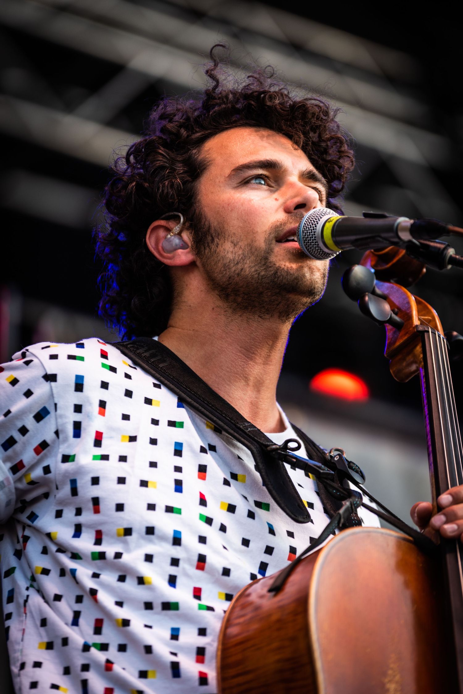 Musician singing into microphone while playing cello in polka dot shirt during outdoor concert.