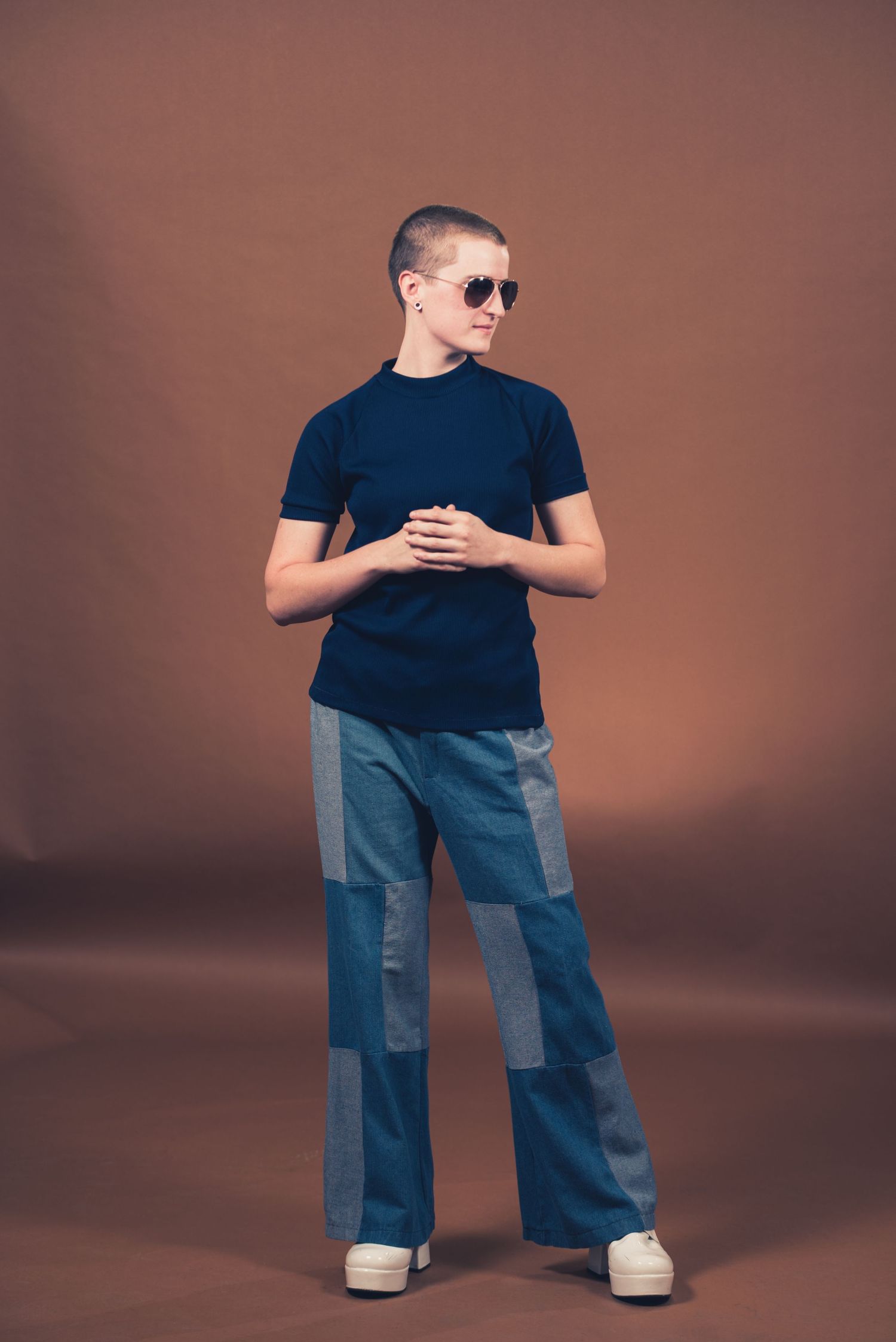 Person in navy t-shirt and patchwork jeans posing with sunglasses against brown backdrop.