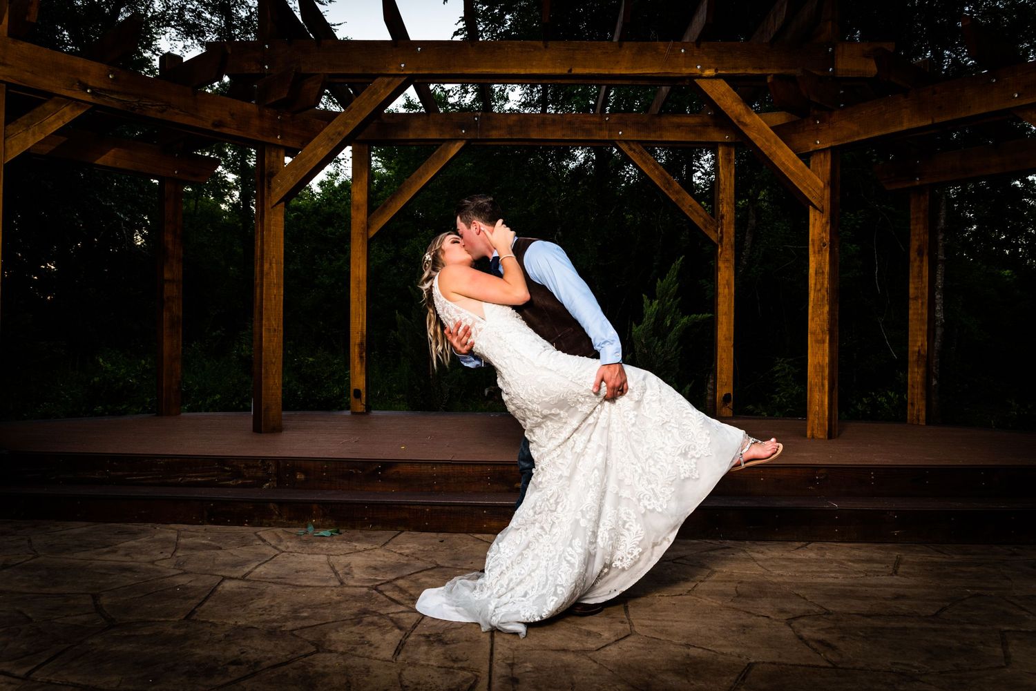 A romantic wedding photo shows a couple in an intimate dip pose under a wooden pavilion at dusk.