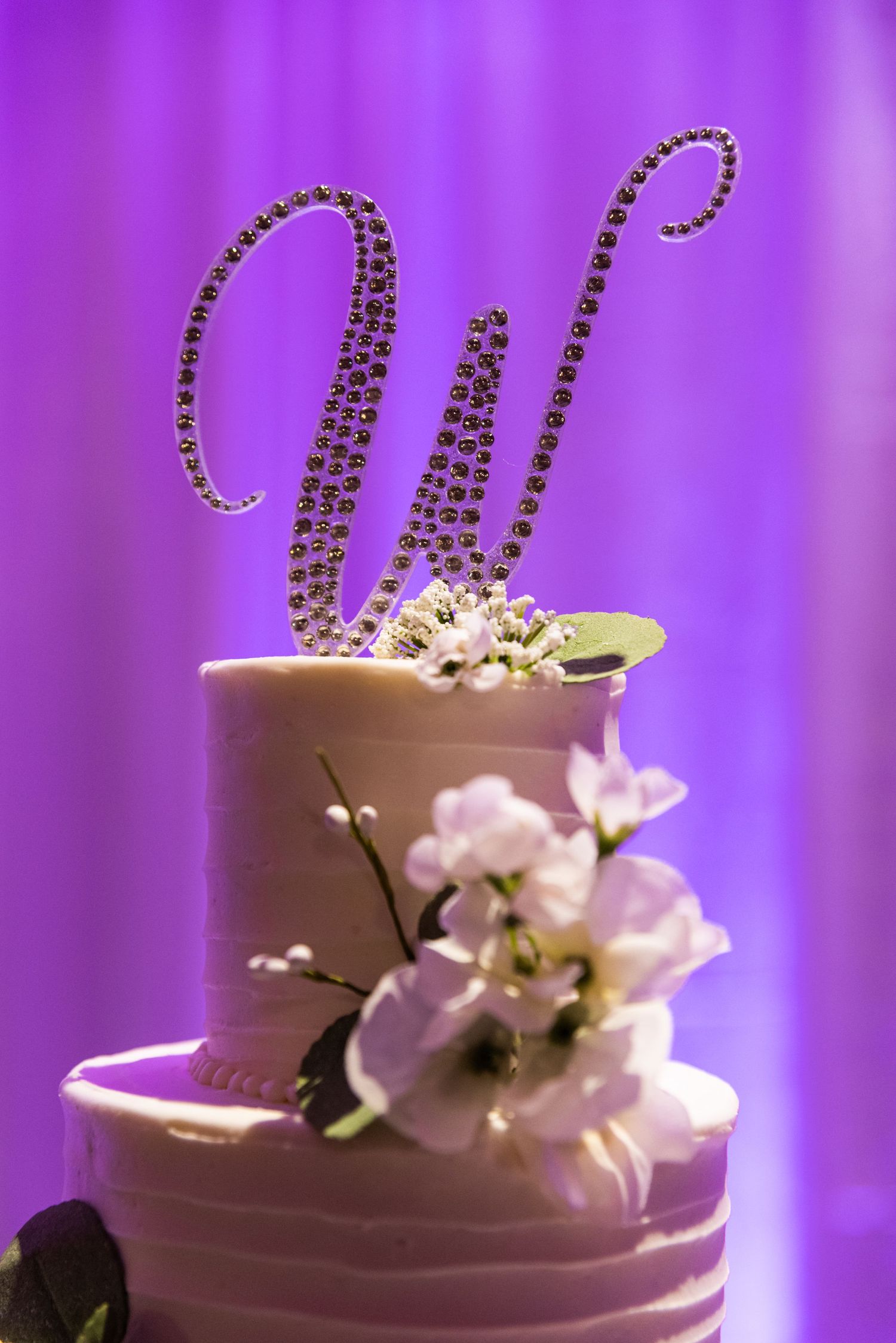 Wedding cake topper featuring a sparkly letter W with purple lighting and white floral decorations.