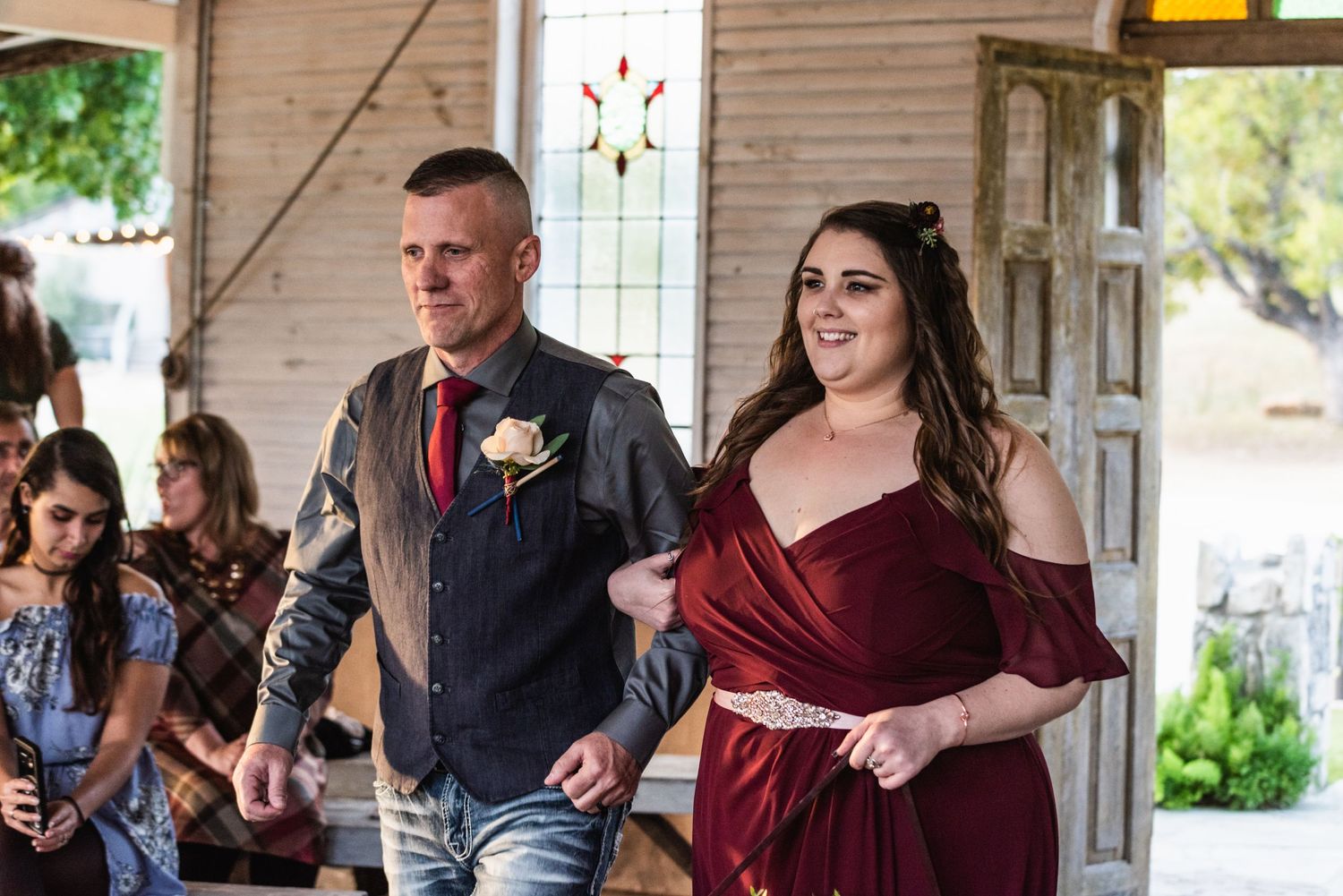 Wedding party members walking down an aisle in burgundy and grey formal attire.