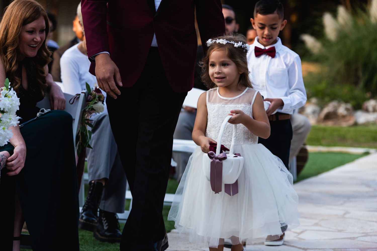Young flower girl in white dress walks down outdoor wedding aisle.