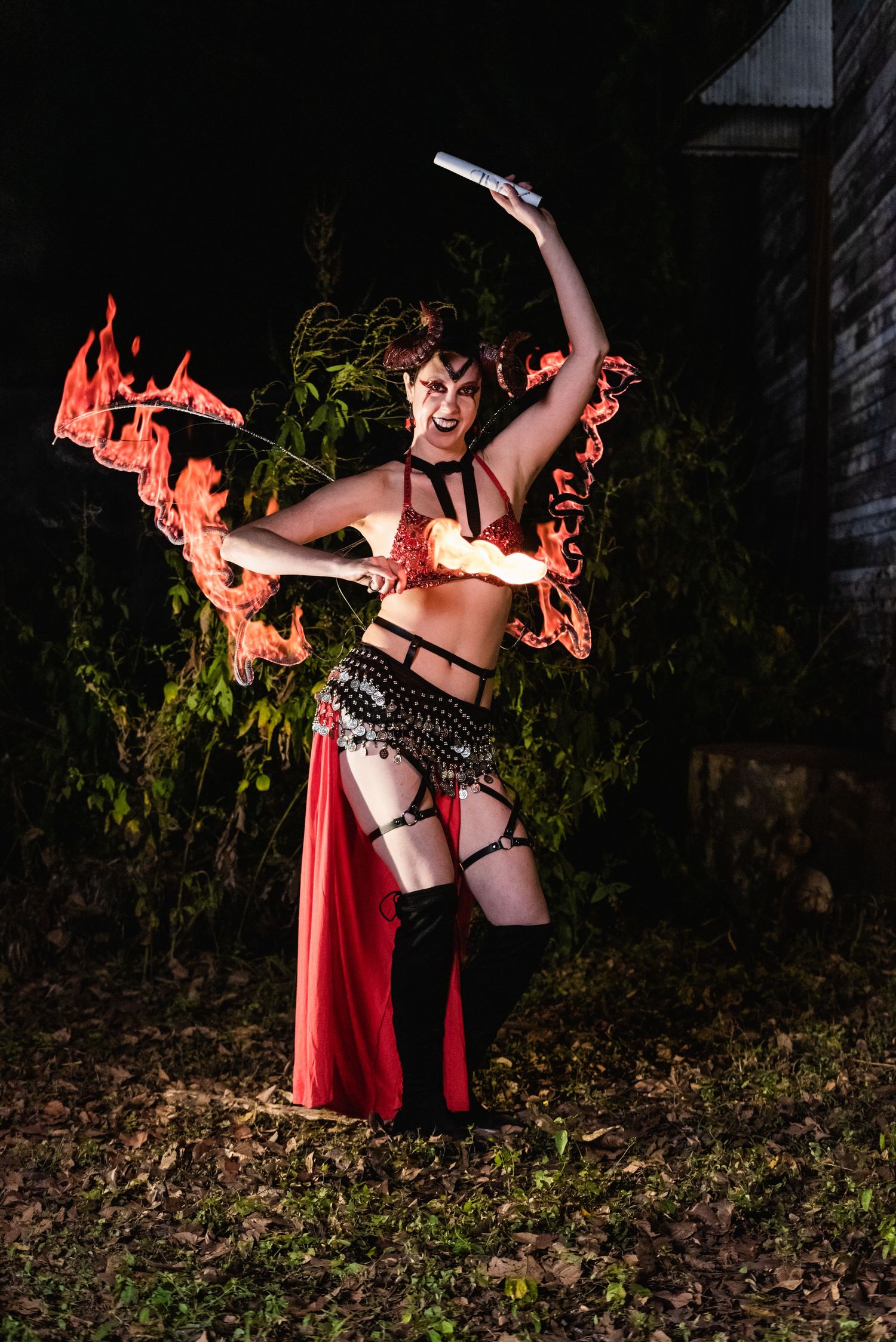 Fire performer in red burlesque costume dances with flaming props outdoors at night.