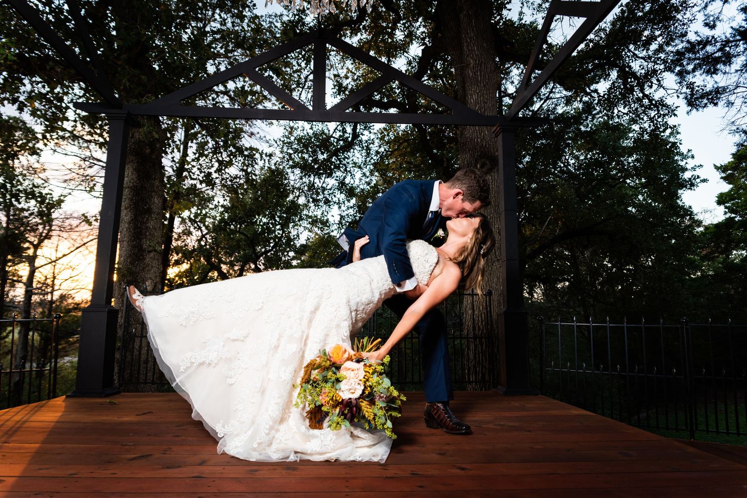 A dramatic dip pose on an outdoor wooden deck with pine trees in the background at sunset.