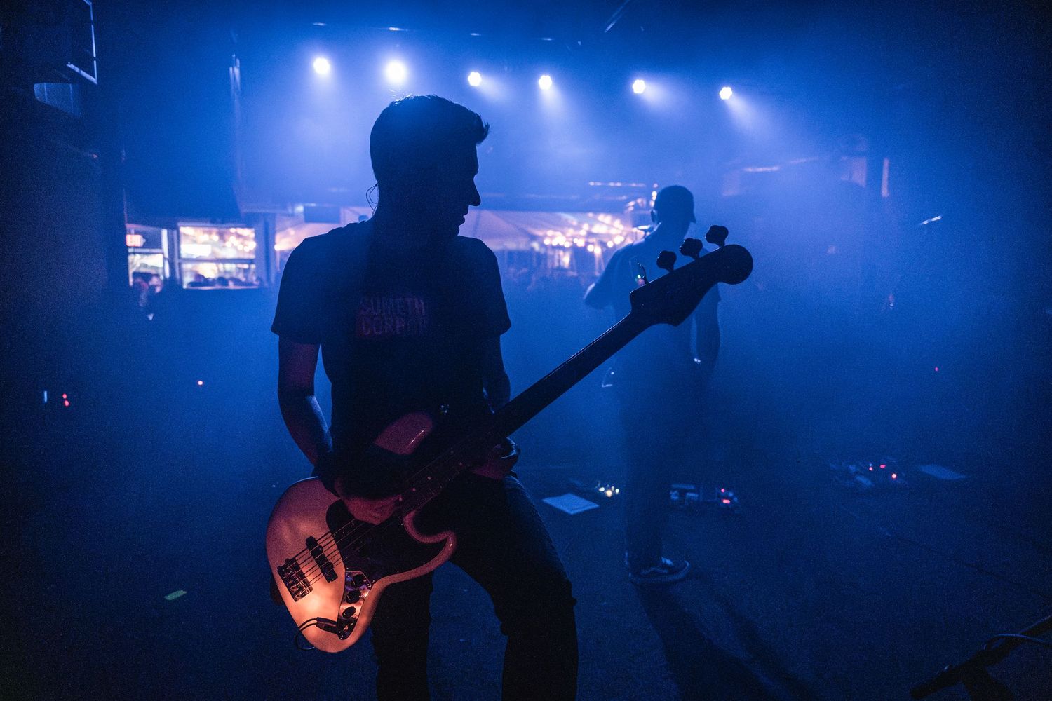 A musician plays bass guitar on a concert stage backlit by blue stage lighting and atmospheric haze.