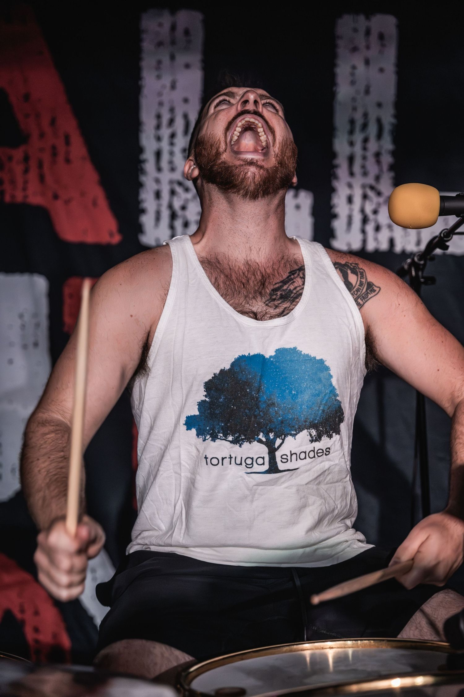 Drummer in white tank top performs with intense emotion during a live concert.