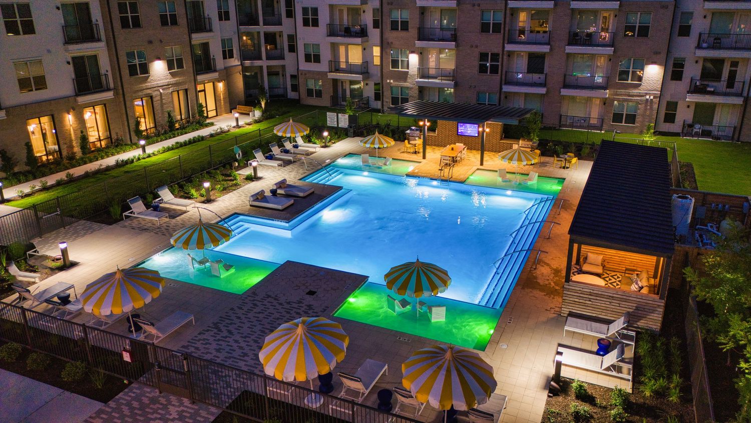Illuminated swimming pool with geometric design surrounded by lounge chairs and apartment buildings at dusk.
