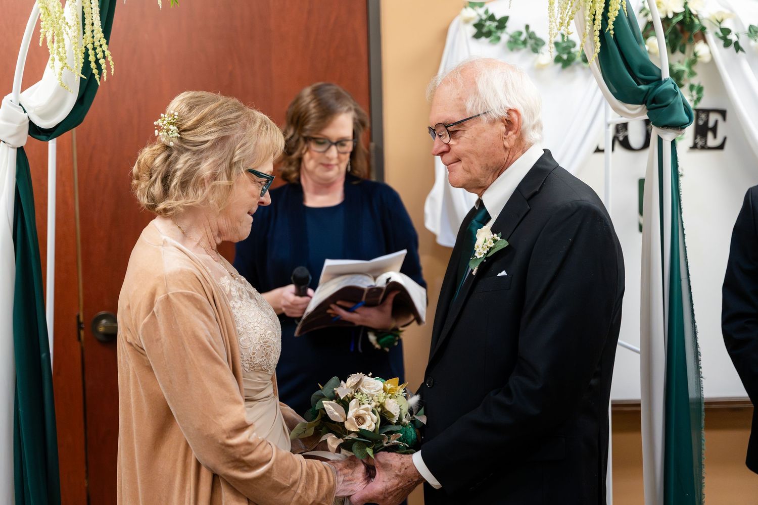 Two people hold hands during a wedding ceremony while an officiant reads from a book.