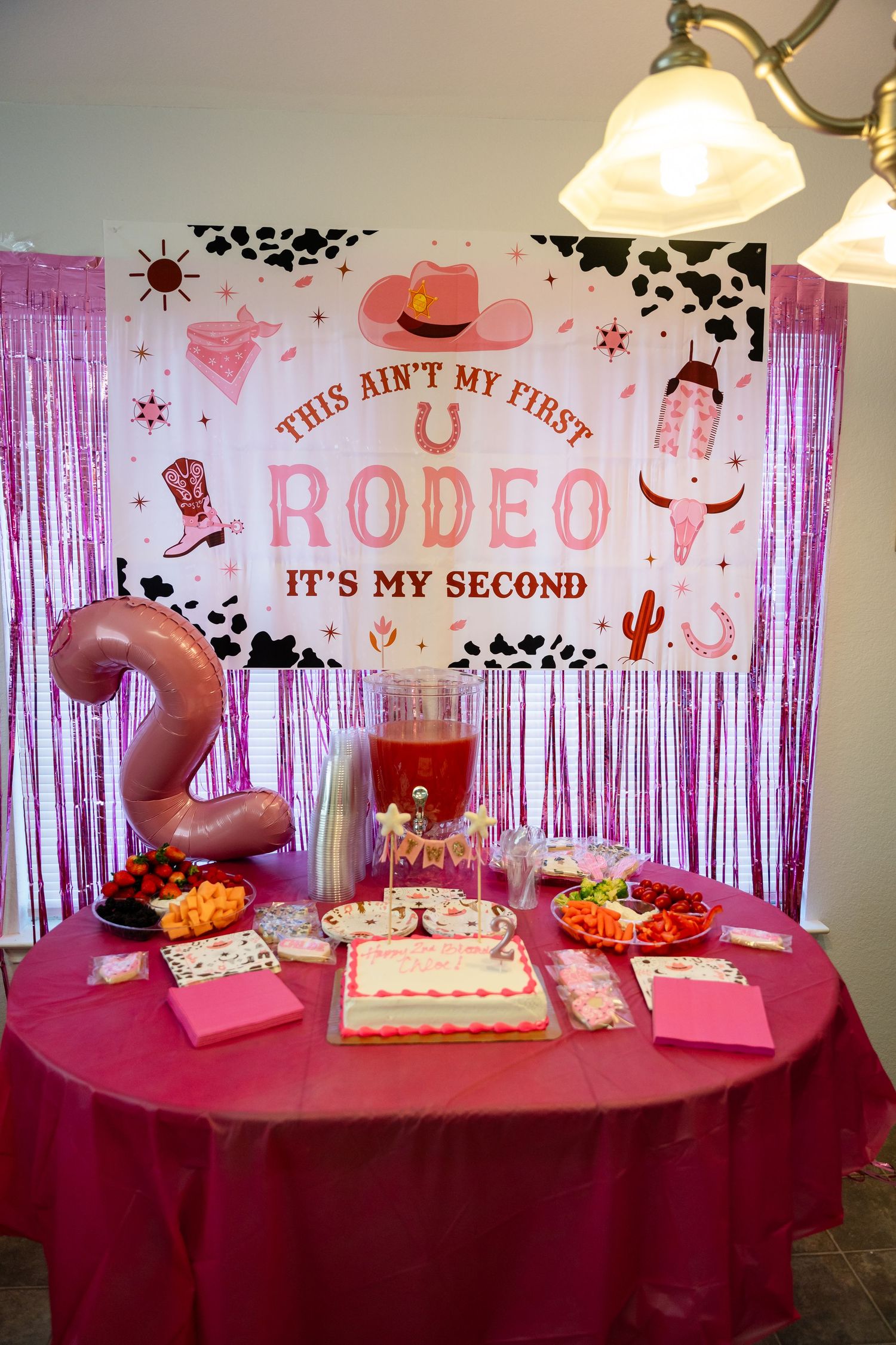 Pink rodeo-themed party table setup with decorations, cake, and snacks for a second birthday celebration.