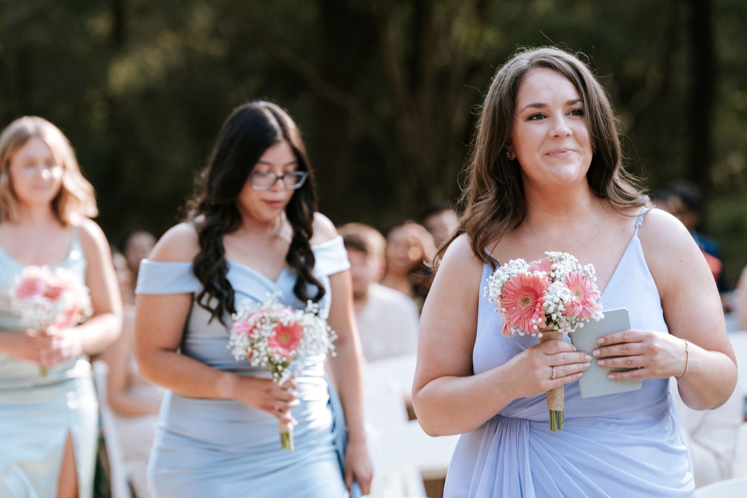 Bridesmaids in light blue dresses carrying pink bouquets during an outdoor ceremony.