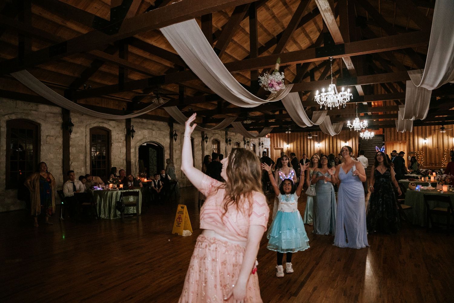A guest in a pink dress dances in a rustic venue with elegant chandeliers and white draping.