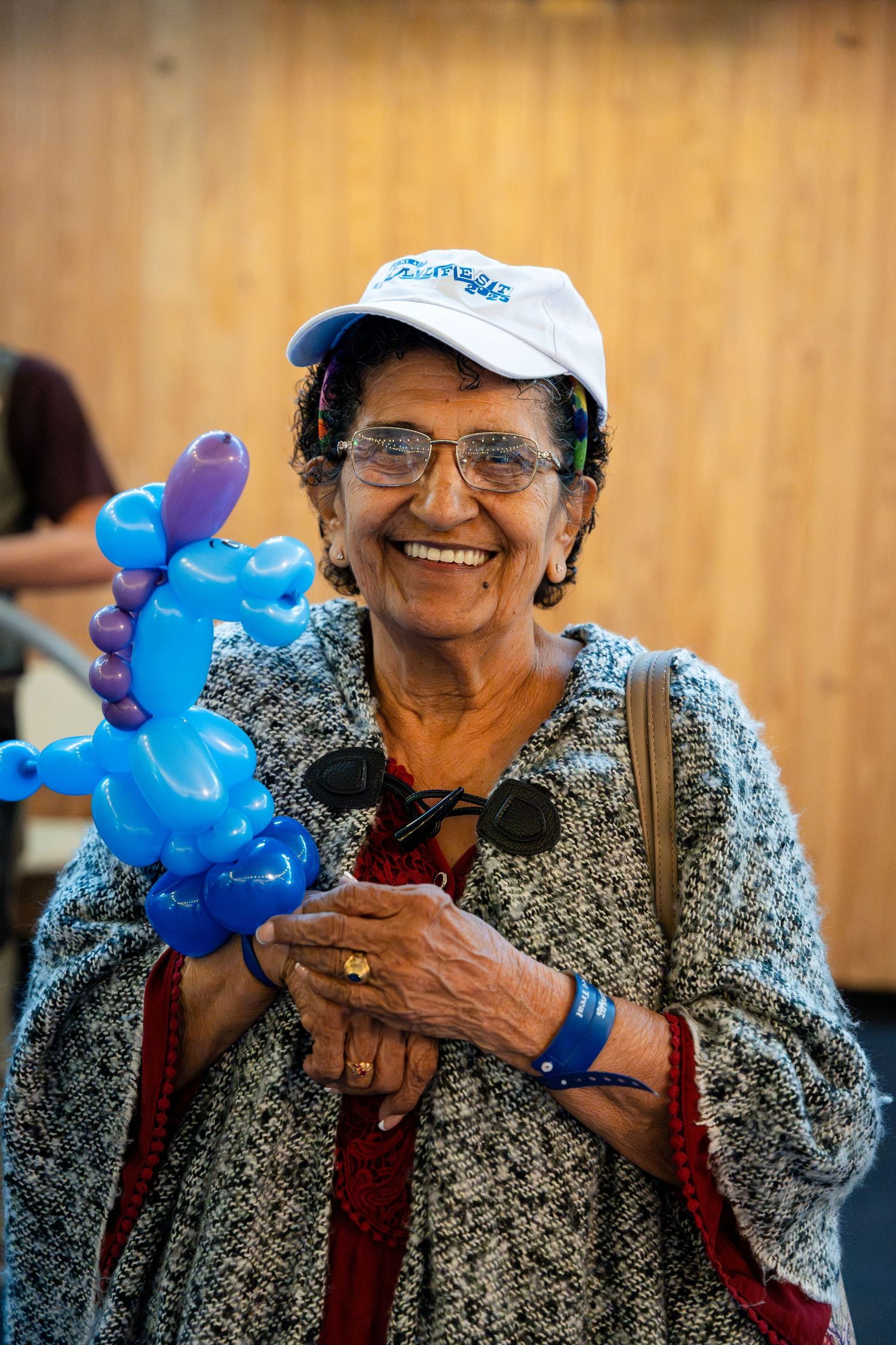 A person wearing glasses and a baseball cap holds a blue balloon animal while smiling outdoors.