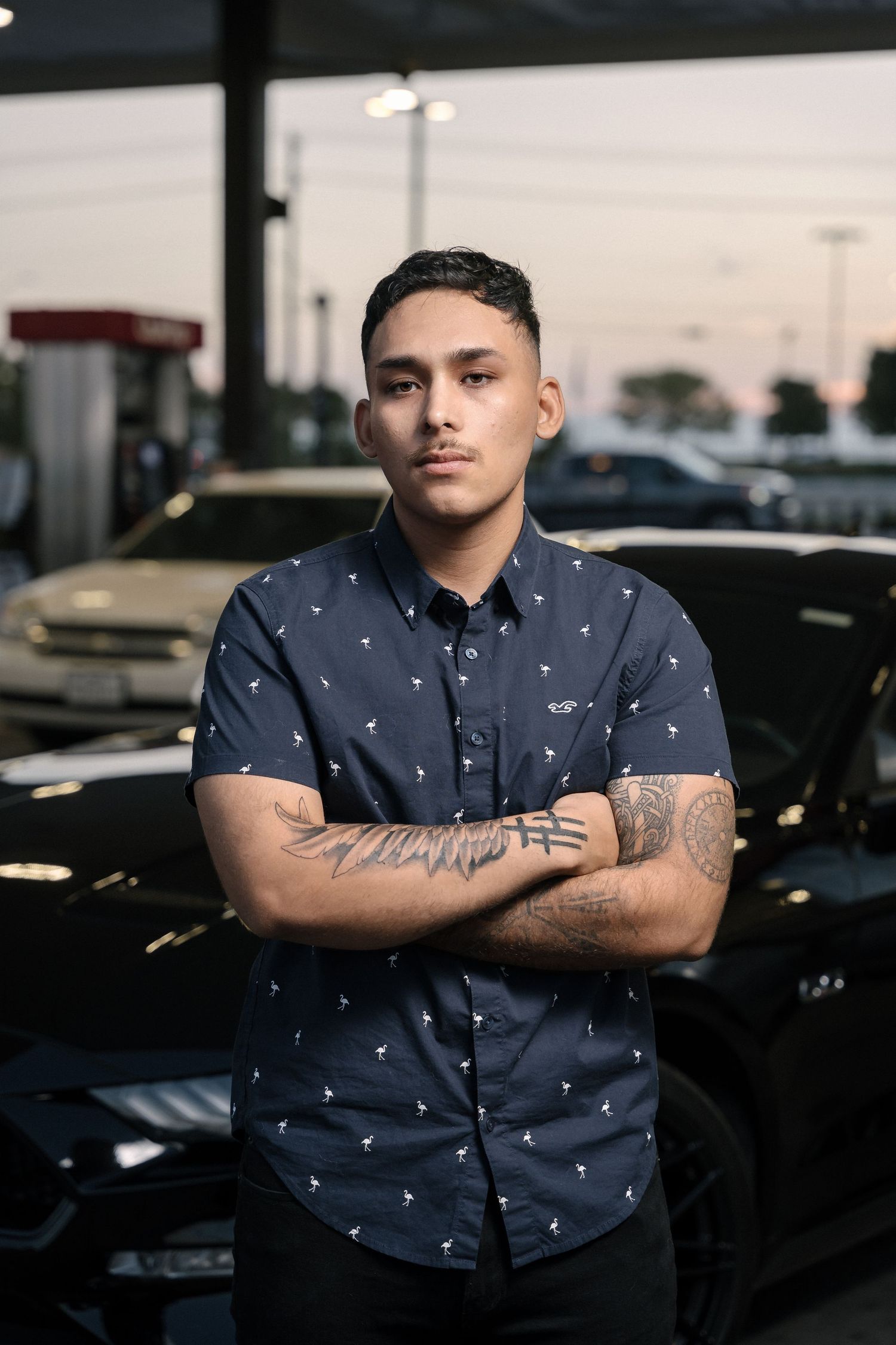 Tattooed person in polka dot shirt posing with crossed arms at dusk near parked cars.