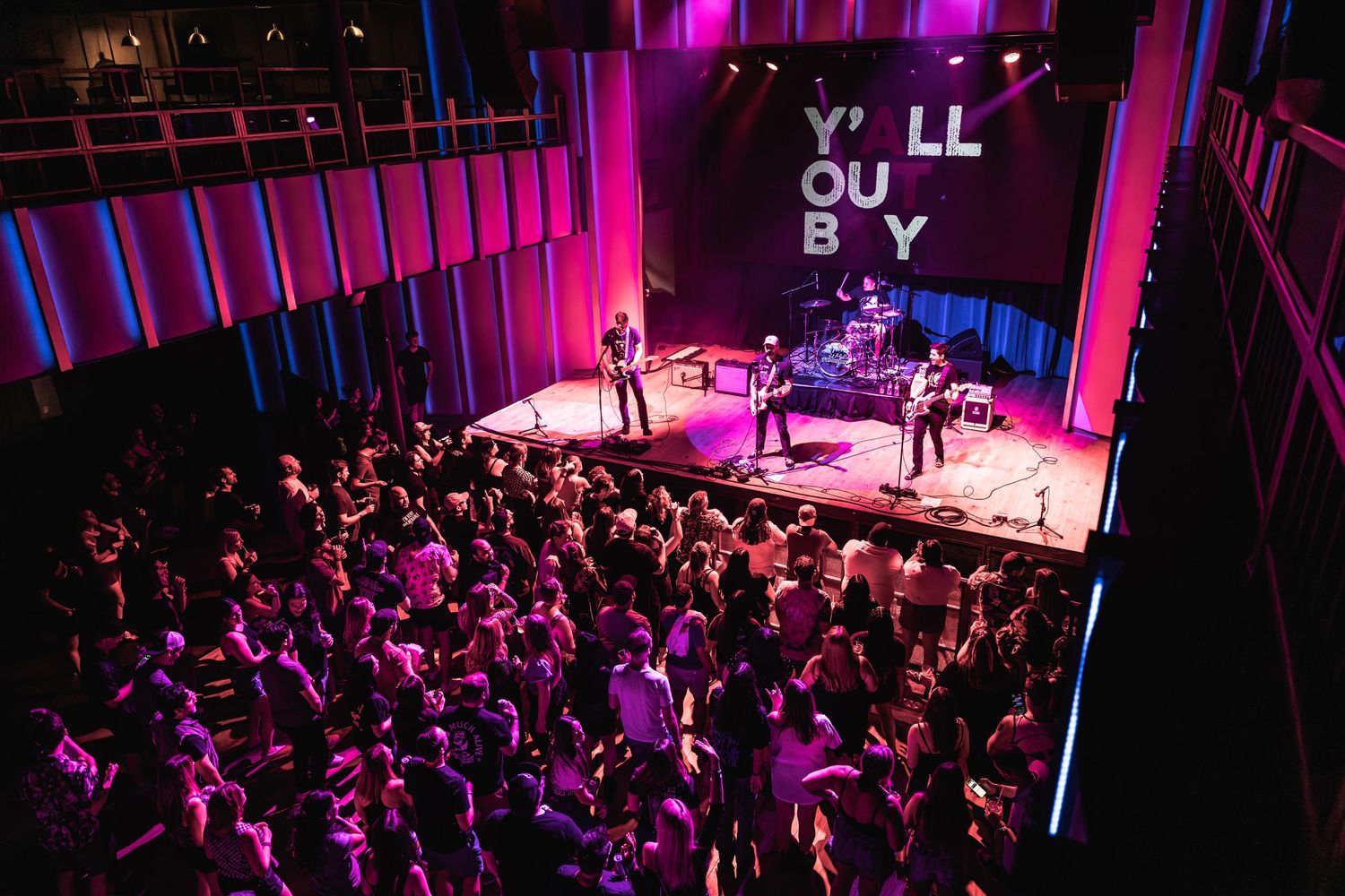 Energetic concert crowd fills venue illuminated by purple lights below 'YOU'LL BE OK' sign on stage.