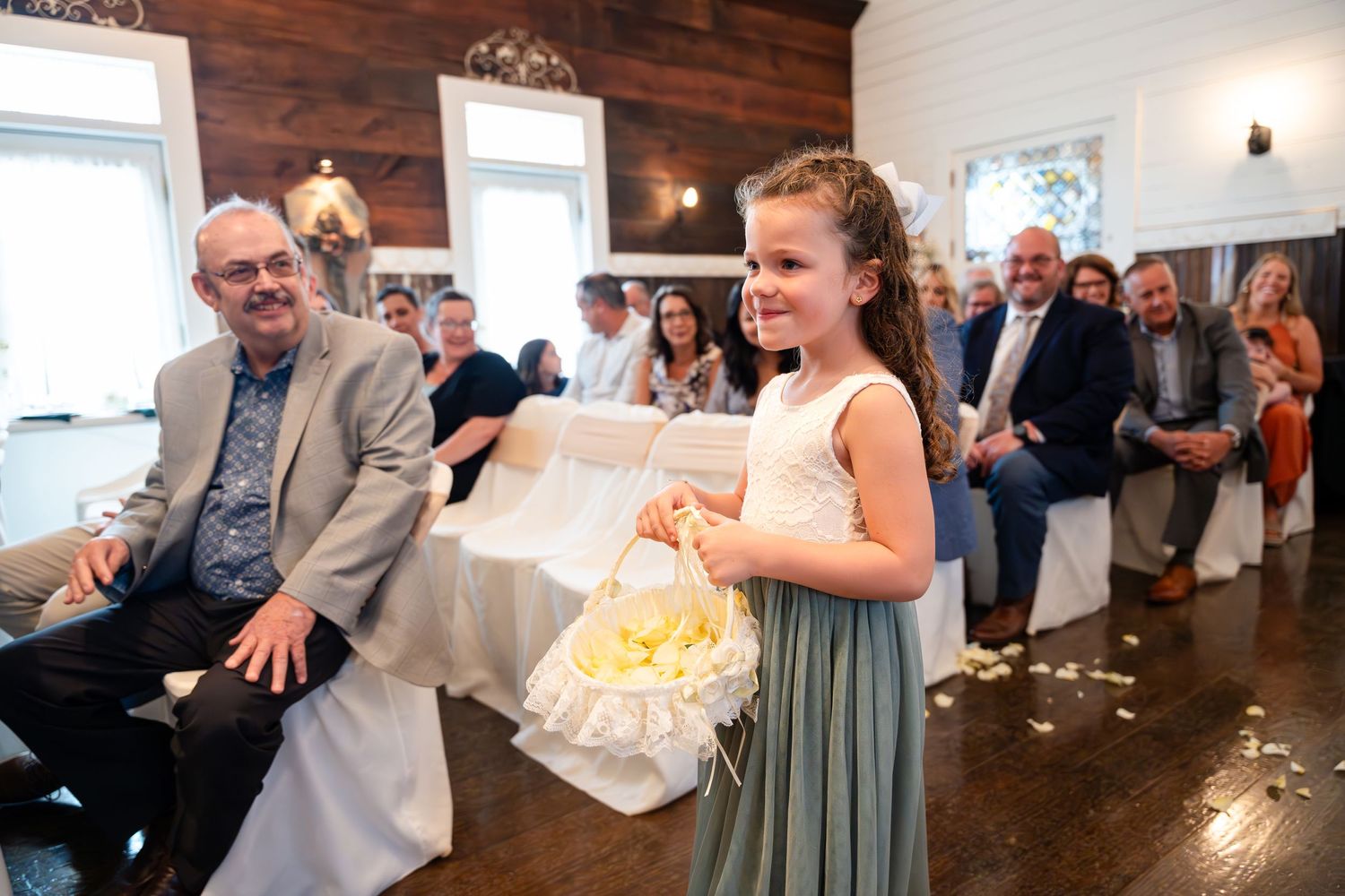 Young flower girl in sage dress carrying petals during wedding ceremony.