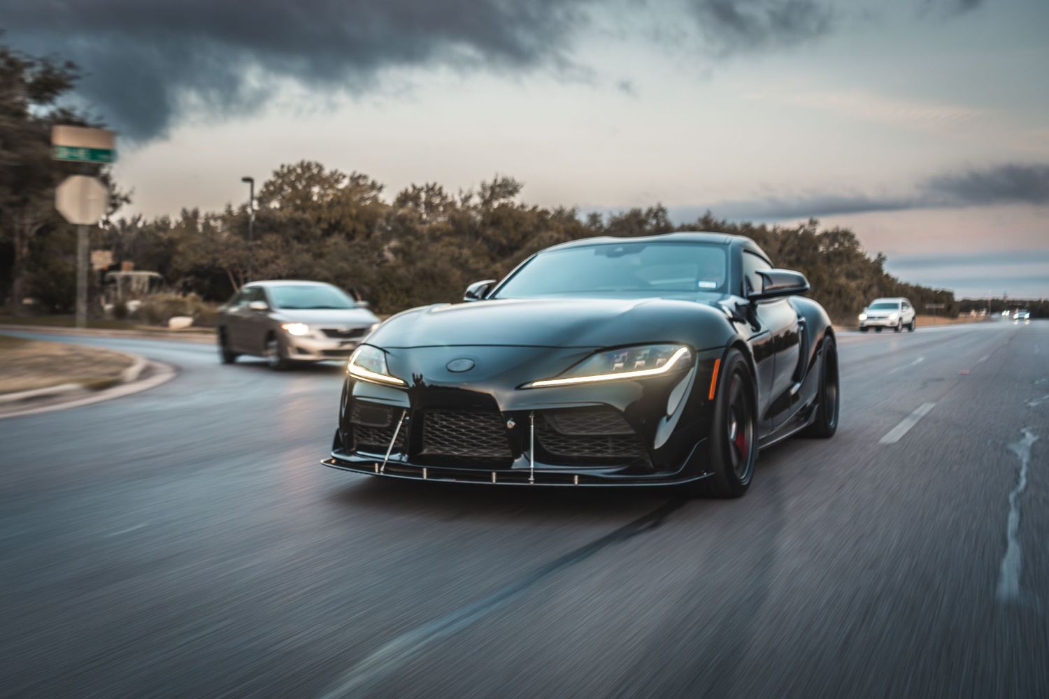 Modified black Toyota Supra with aggressive body kit driving on a cloudy day.
