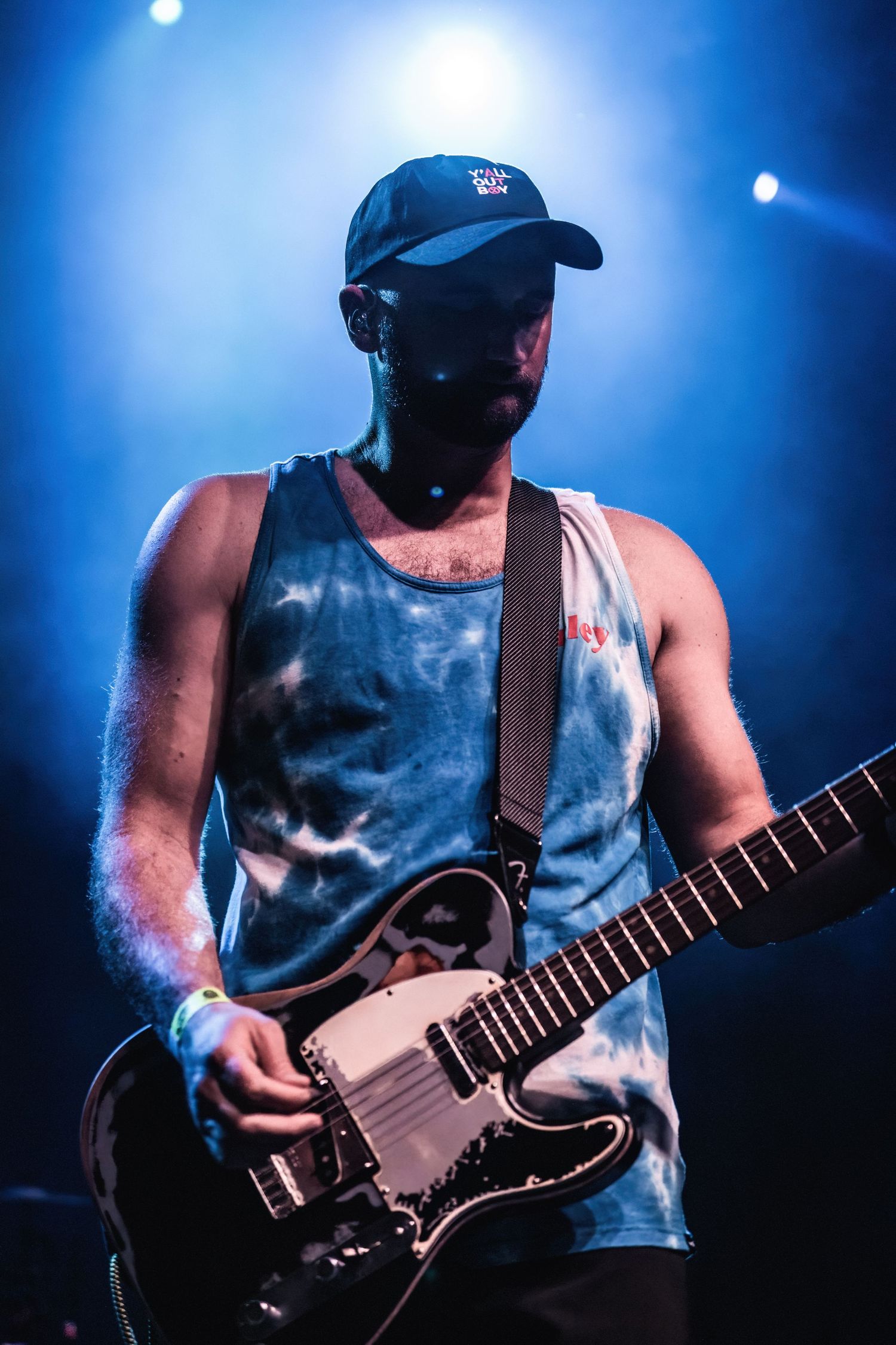 Guitar player in blue cap and tie-dye tank top performs under dramatic stage lighting.