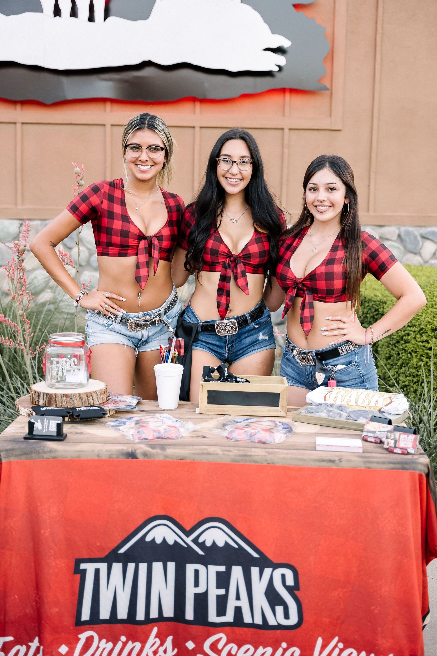 Three staff members in matching plaid tops at Twin Peaks promotional booth with merchandise display.