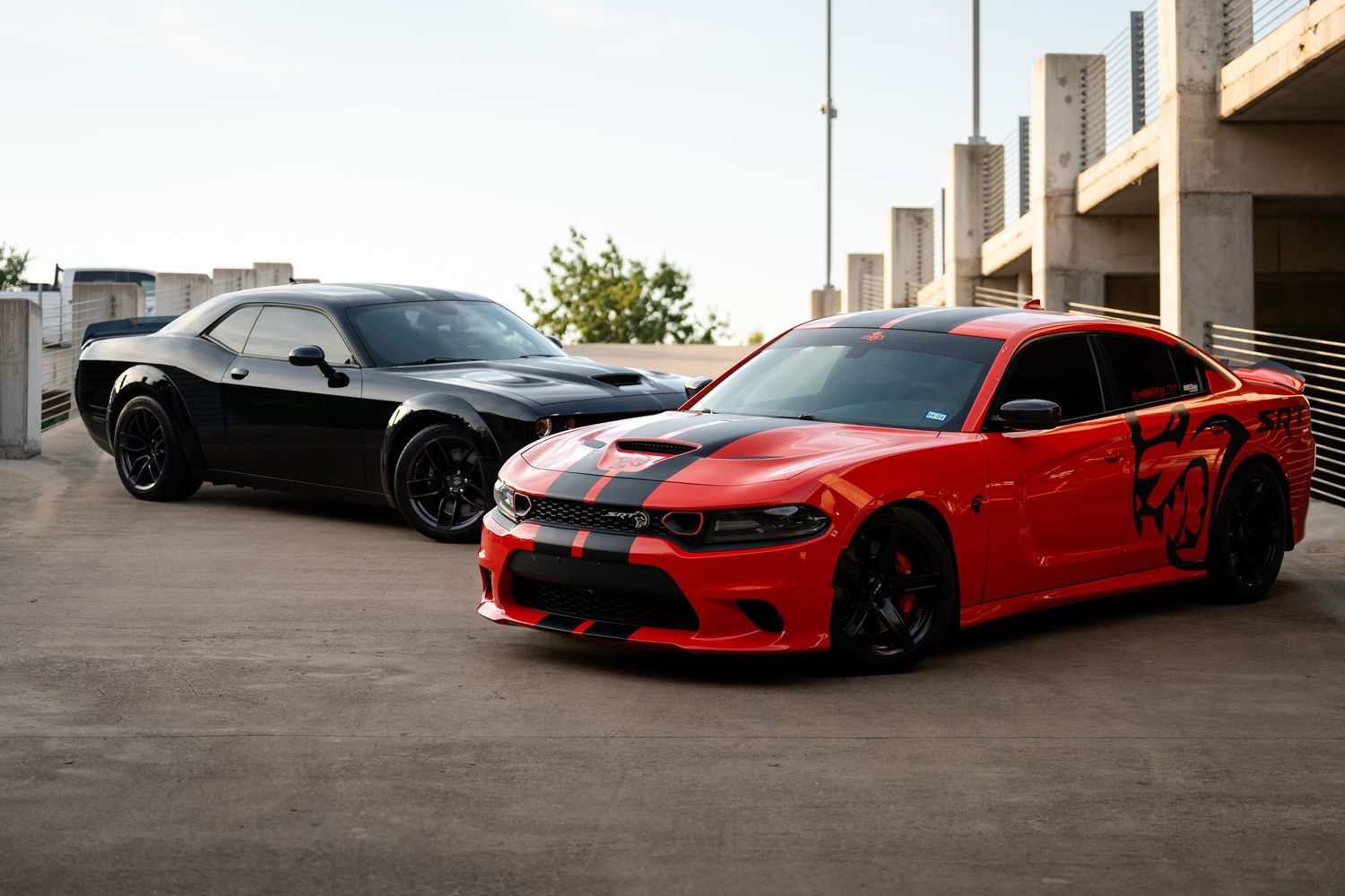 A black Dodge Challenger and red Dodge Charger SRT with custom modifications parked in a concrete structure.