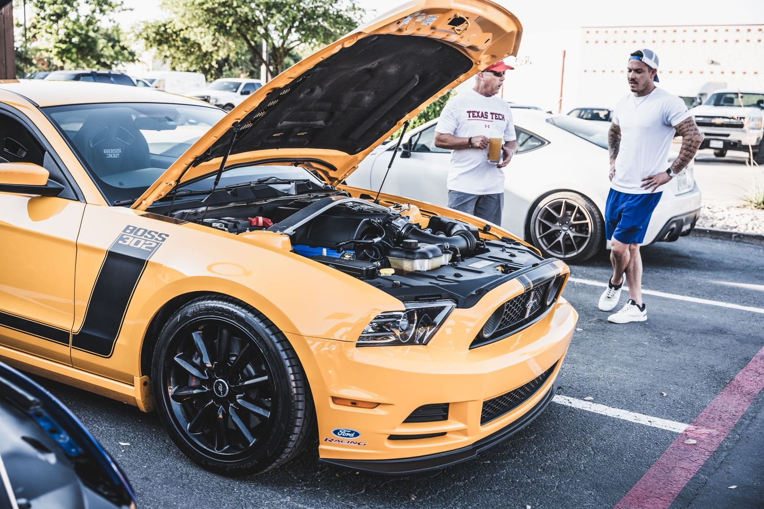 Custom orange Mustang with hood open at a car show in a parking lot.