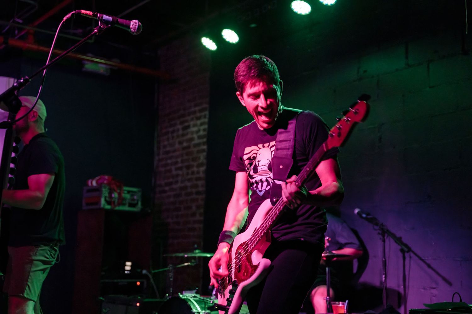Musician playing bass guitar under purple and green stage lighting in a brick-walled venue.