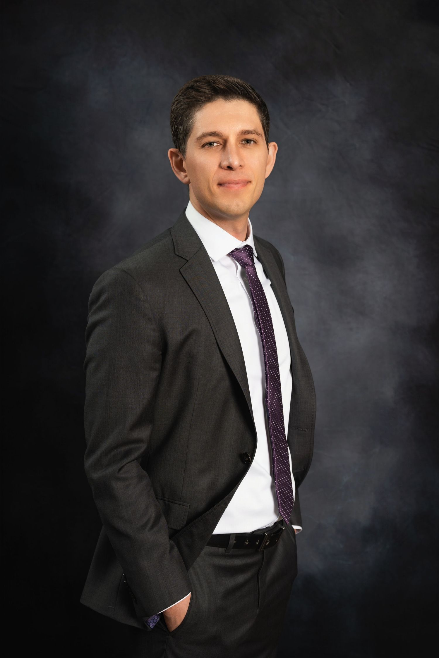 Formal business portrait with hand in pocket pose against textured dark background.