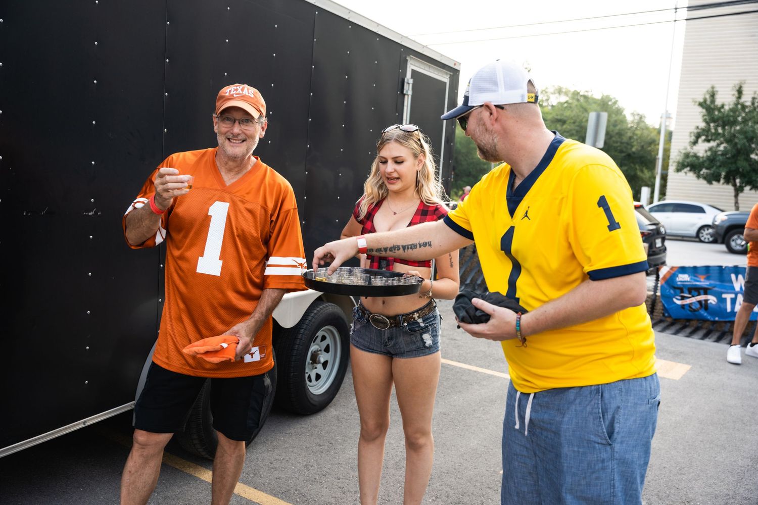 Three sports fans in team jerseys share drinks near a black trailer at a tailgate party.
