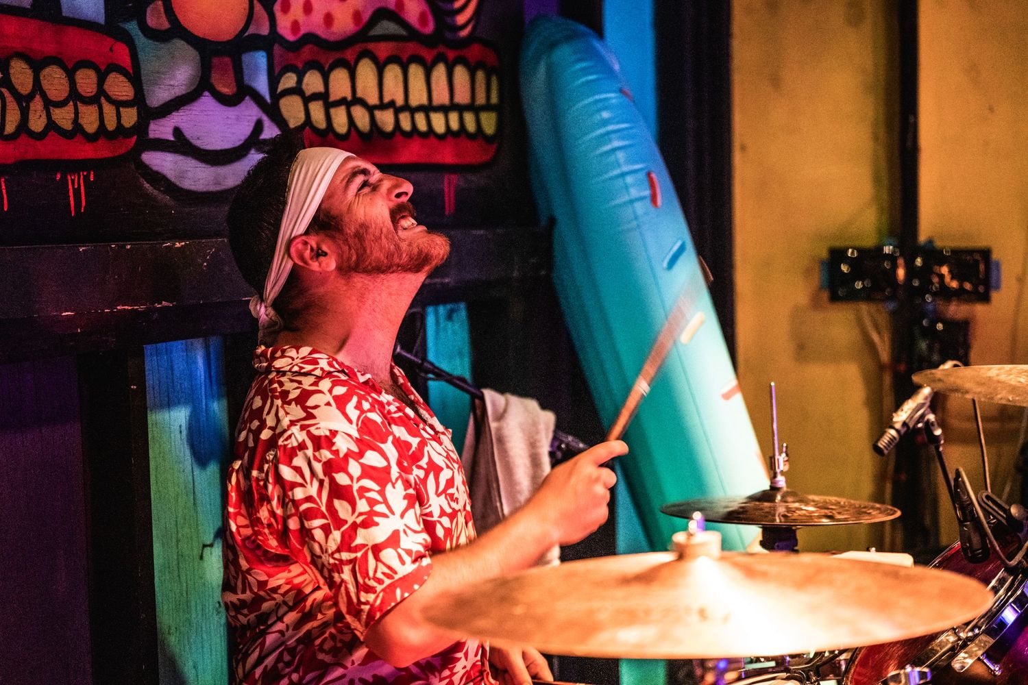 Energetic drummer performs in a red Hawaiian shirt at a vibrant music venue.