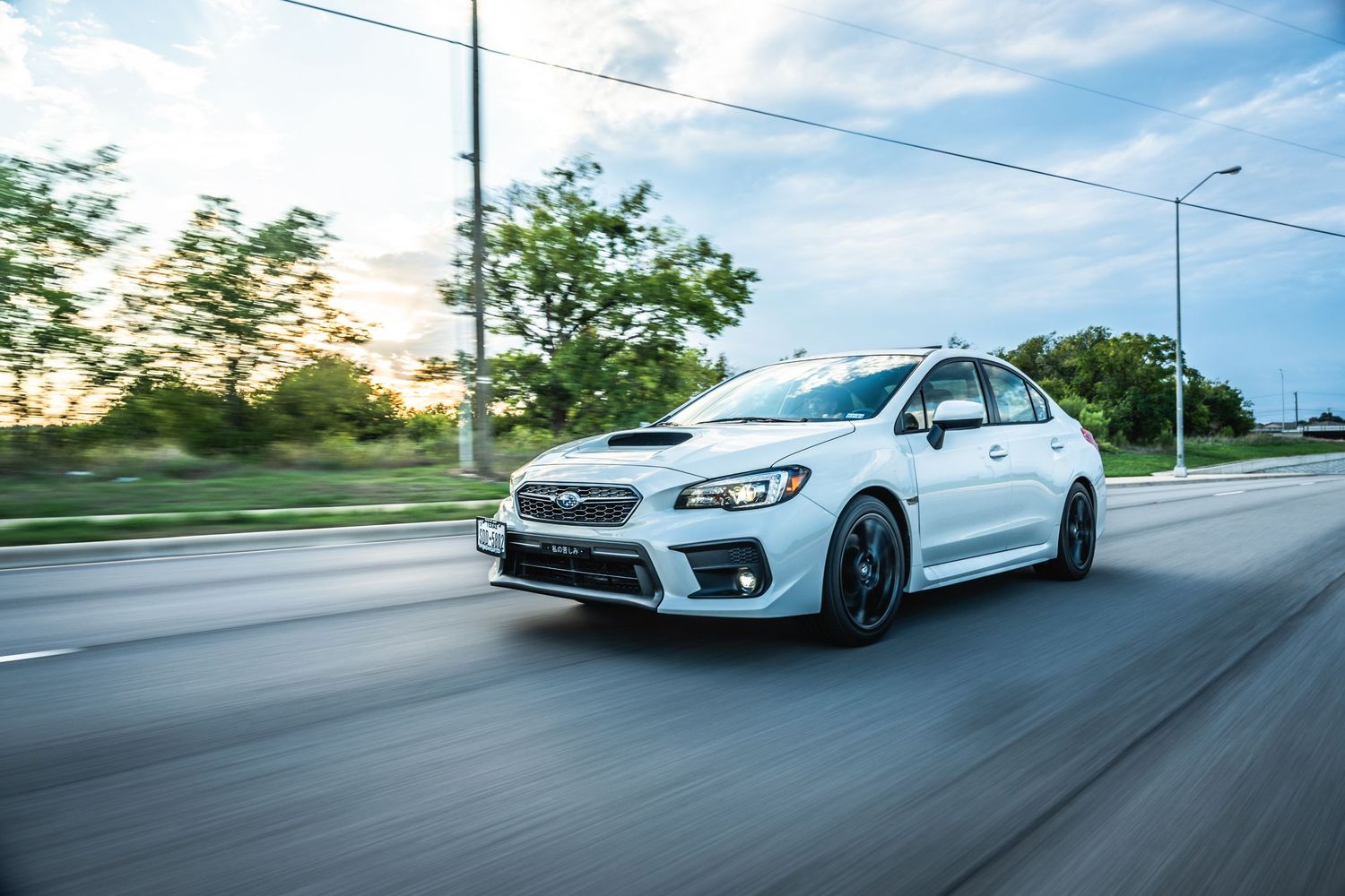 Pearl white Subaru WRX with aftermarket wheels captured in motion during golden hour.