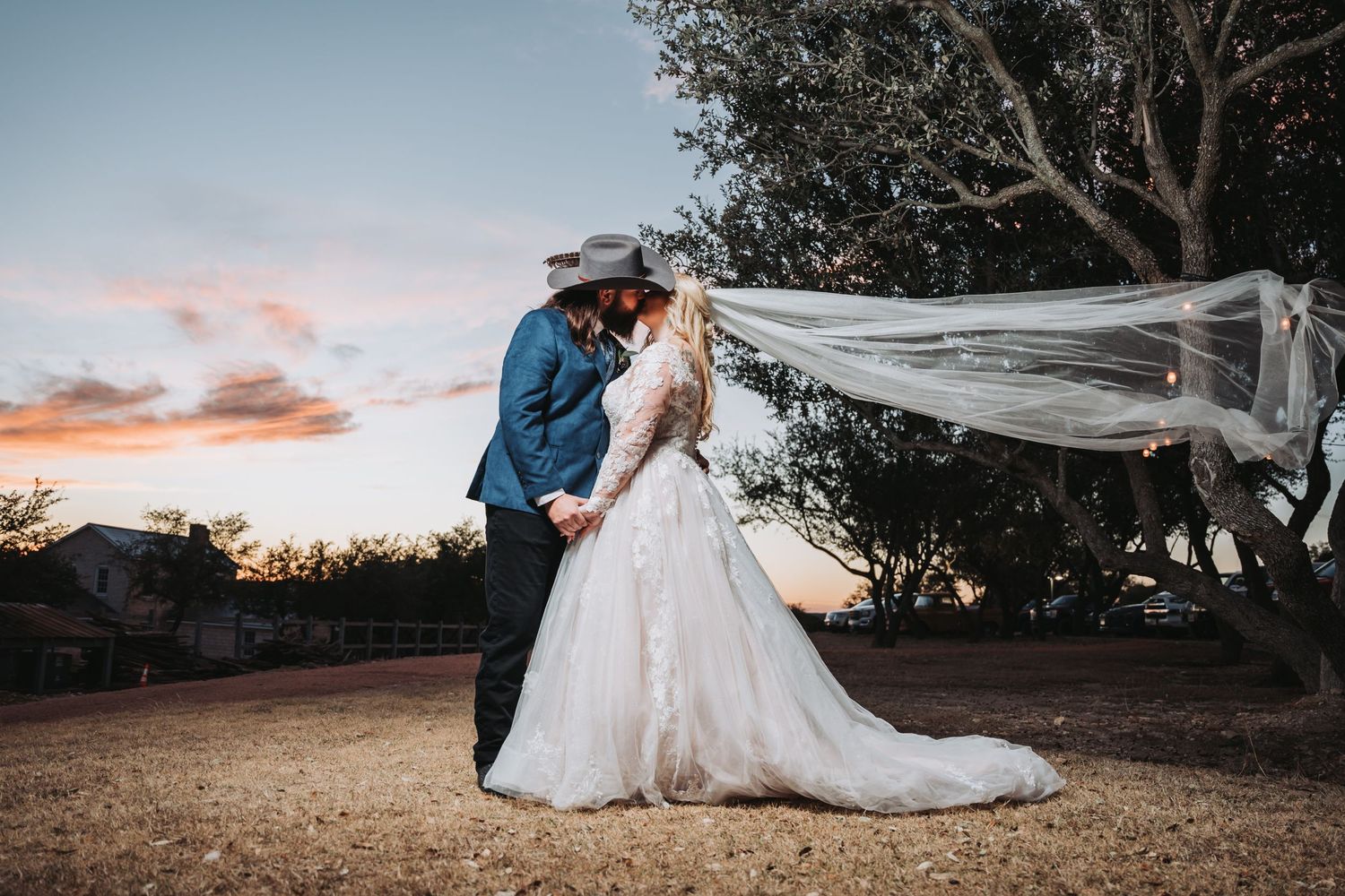 A dramatic sunset wedding photo with the bride's veil flowing in the wind during an outdoor ceremony.