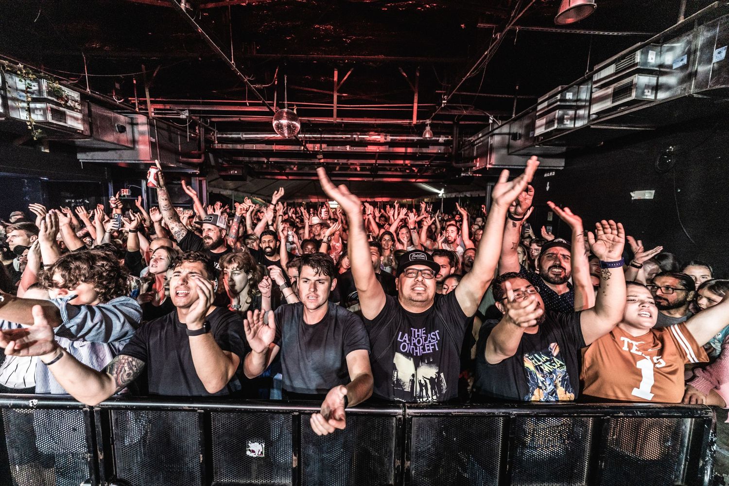 Packed concert venue crowd raises hands in unison during energetic performance.